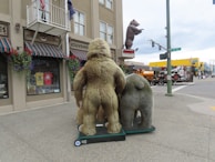 A street scene featuring large bear statues outside a souvenir store with striped canopies. The bears are mounted on wheeled platforms facing the street corner. There's a large bear sculpture in the distance atop a building. Various flags are displayed, and pedestrians are visible in the background.