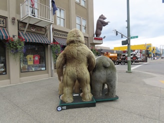 A street scene featuring large bear statues outside a souvenir store with striped canopies. The bears are mounted on wheeled platforms facing the street corner. There's a large bear sculpture in the distance atop a building. Various flags are displayed, and pedestrians are visible in the background.