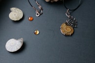 A collection of jewelry pieces is displayed on a textured dark background, including a pendant with a fossil-like spiral design, an earring with an empty hook, and several round amber-colored beads.