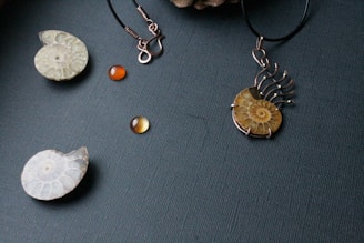 A collection of jewelry pieces is displayed on a textured dark background, including a pendant with a fossil-like spiral design, an earring with an empty hook, and several round amber-colored beads.