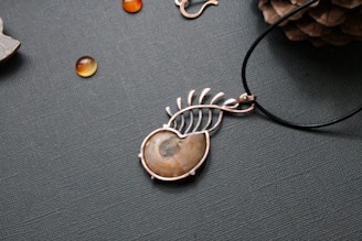 A piece of jewelry with a gemstone set in a silver wire wrapping design that resembles a leaf or feather. Nearby, small polished stones or beads are scattered on a textured surface, and a pine cone is partially visible in the upper right corner.