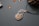 A piece of jewelry with a gemstone set in a silver wire wrapping design that resembles a leaf or feather. Nearby, small polished stones or beads are scattered on a textured surface, and a pine cone is partially visible in the upper right corner.