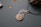 A piece of jewelry with a gemstone set in a silver wire wrapping design that resembles a leaf or feather. Nearby, small polished stones or beads are scattered on a textured surface, and a pine cone is partially visible in the upper right corner.