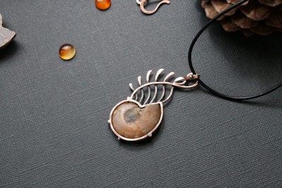 A piece of jewelry with a gemstone set in a silver wire wrapping design that resembles a leaf or feather. Nearby, small polished stones or beads are scattered on a textured surface, and a pine cone is partially visible in the upper right corner.