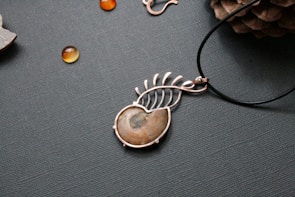 A piece of jewelry with a gemstone set in a silver wire wrapping design that resembles a leaf or feather. Nearby, small polished stones or beads are scattered on a textured surface, and a pine cone is partially visible in the upper right corner.