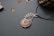 A piece of jewelry with a gemstone set in a silver wire wrapping design that resembles a leaf or feather. Nearby, small polished stones or beads are scattered on a textured surface, and a pine cone is partially visible in the upper right corner.