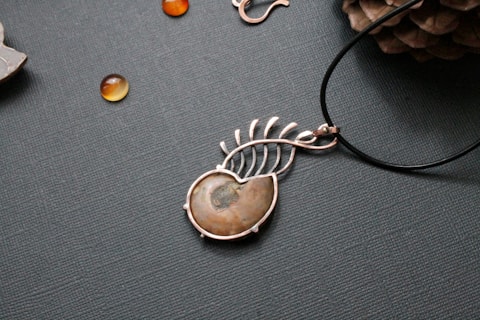 A piece of jewelry with a gemstone set in a silver wire wrapping design that resembles a leaf or feather. Nearby, small polished stones or beads are scattered on a textured surface, and a pine cone is partially visible in the upper right corner.