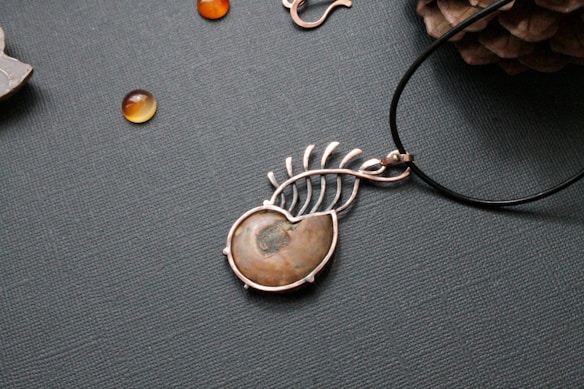 A piece of jewelry with a gemstone set in a silver wire wrapping design that resembles a leaf or feather. Nearby, small polished stones or beads are scattered on a textured surface, and a pine cone is partially visible in the upper right corner.