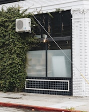 A building facade featuring a white air conditioning unit mounted on the exterior wall, partially covered with creeping green ivy. The building is painted white with black-tiled lower sections, and an industrial light fixture is attached above a large window with frosted glass. The sidewalk in front is made of concrete with a red-painted curb.