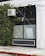 A building facade featuring a white air conditioning unit mounted on the exterior wall, partially covered with creeping green ivy. The building is painted white with black-tiled lower sections, and an industrial light fixture is attached above a large window with frosted glass. The sidewalk in front is made of concrete with a red-painted curb.