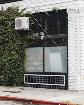 A building facade featuring a white air conditioning unit mounted on the exterior wall, partially covered with creeping green ivy. The building is painted white with black-tiled lower sections, and an industrial light fixture is attached above a large window with frosted glass. The sidewalk in front is made of concrete with a red-painted curb.