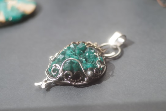 A silver pendant with intricate wirework encases a vibrant green stone, displaying an ornate design. The pendant lies on a gray surface, casting a soft shadow.