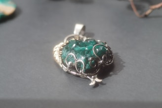 A green gemstone pendant with intricate silver wire wrapping is placed on a smooth, grey surface. The stone has a rough texture and is surrounded by silver loops forming an artistic design. Soft light illuminates the pendant, casting a gentle shadow.