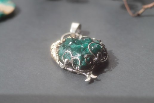 A green gemstone pendant with intricate silver wire wrapping is placed on a smooth, grey surface. The stone has a rough texture and is surrounded by silver loops forming an artistic design. Soft light illuminates the pendant, casting a gentle shadow.