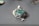 A green gemstone pendant with intricate silver wire wrapping is placed on a smooth, grey surface. The stone has a rough texture and is surrounded by silver loops forming an artistic design. Soft light illuminates the pendant, casting a gentle shadow.