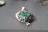 A green gemstone pendant with intricate silver wire wrapping is placed on a smooth, grey surface. The stone has a rough texture and is surrounded by silver loops forming an artistic design. Soft light illuminates the pendant, casting a gentle shadow.
