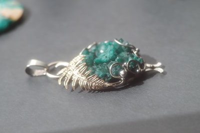 A silver pendant with intricate wirework encases a rough, textured green stone, lying on a smooth, gray surface. The metal appears intricately twisted and woven, creating a decorative and elegant design around the central gemstone.