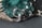 A close-up view of an intricately designed piece of jewelry featuring a large, raw green gemstone. The gemstone is encased in a silver-colored metal setting with swirling patterns and small decorative silver balls.