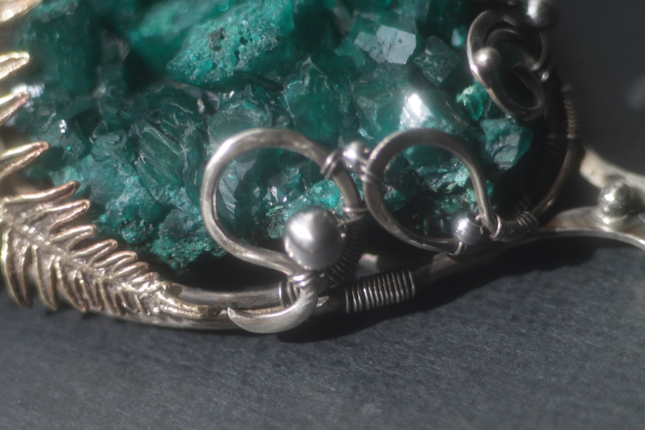A close-up view of an intricately designed piece of jewelry featuring a large, raw green gemstone. The gemstone is encased in a silver-colored metal setting with swirling patterns and small decorative silver balls.