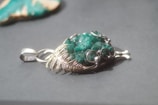 A silver pendant featuring a textured green stone is displayed on a smooth gray surface. The pendant has intricate metalwork resembling leaf patterns, securely wrapping around the stone, which has a rough, unpolished appearance. The background includes a blurred object, possibly another stone or piece of jewelry, suggesting a setting focused on accessories.
