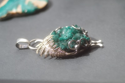 A silver pendant featuring a textured green stone is displayed on a smooth gray surface. The pendant has intricate metalwork resembling leaf patterns, securely wrapping around the stone, which has a rough, unpolished appearance. The background includes a blurred object, possibly another stone or piece of jewelry, suggesting a setting focused on accessories.