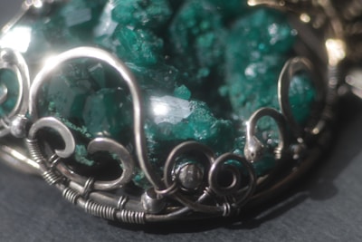 An intricate piece of jewelry featuring a cluster of raw, emerald-green crystals set in an ornate silver frame with swirling metalwork design.