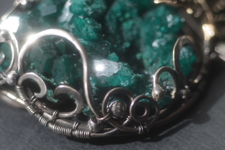 An intricate piece of jewelry featuring a cluster of raw, emerald-green crystals set in an ornate silver frame with swirling metalwork design.