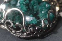 An intricate piece of jewelry featuring a cluster of raw, emerald-green crystals set in an ornate silver frame with swirling metalwork design.