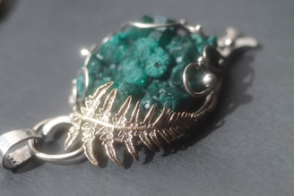 An elegant piece of jewelry featuring a vibrant green, textured gemstone encased in intricate silver metalwork with a leaf motif. The pendant is set against a dark, neutral background, highlighting the gemstone's color and the metallic details.