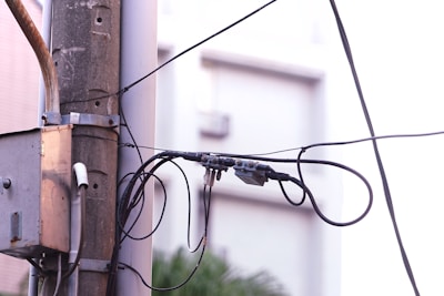A utility pole is depicted with multiple black wires and cables attached. A grey electrical junction box is mounted on the pole. The background appears to be an out-of-focus building with a light, soft color palette. The setting seems urban and the focus is on the interconnected network of cables.
