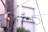 A utility pole is depicted with multiple black wires and cables attached. A grey electrical junction box is mounted on the pole. The background appears to be an out-of-focus building with a light, soft color palette. The setting seems urban and the focus is on the interconnected network of cables.