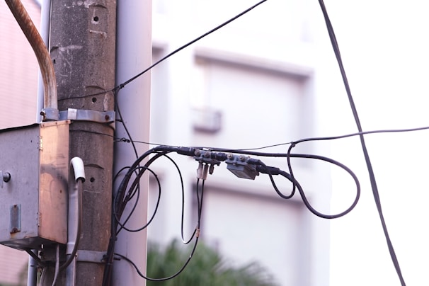 A utility pole is depicted with multiple black wires and cables attached. A grey electrical junction box is mounted on the pole. The background appears to be an out-of-focus building with a light, soft color palette. The setting seems urban and the focus is on the interconnected network of cables.
