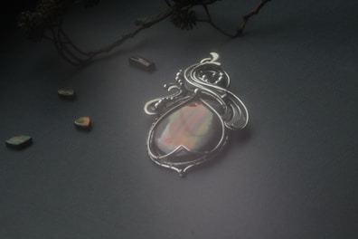 An ornate silver pendant featuring an opalescent stone placed on a dark, textured surface. Surrounding the pendant are small, polished stones that reflect various iridescent colors. A delicate branch with intricate details is visible in the background, adding an organic element to the composition.