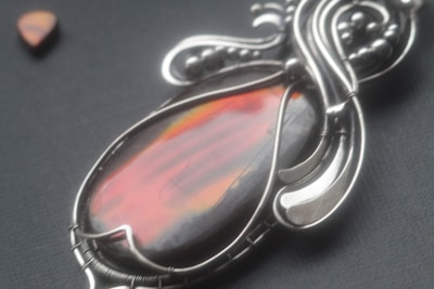 A close-up of an ornate pendant featuring a large, polished stone with vibrant red and orange hues. The stone is set in an intricate metal frame with flowing, decorative designs. In the background, there is a small triangular stone with similar coloring.