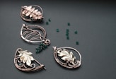 A collection of intricately designed metallic jewelry pieces in various shapes, featuring floral and leaf motifs. The pieces are adorned with small, round, deep green beads scattered on a textured dark surface.