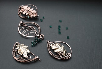 A collection of intricately designed metallic jewelry pieces in various shapes, featuring floral and leaf motifs. The pieces are adorned with small, round, deep green beads scattered on a textured dark surface.