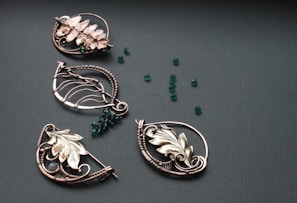 A collection of intricately designed metallic jewelry pieces in various shapes, featuring floral and leaf motifs. The pieces are adorned with small, round, deep green beads scattered on a textured dark surface.