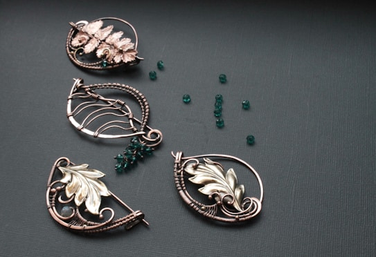 A collection of intricately designed metallic jewelry pieces in various shapes, featuring floral and leaf motifs. The pieces are adorned with small, round, deep green beads scattered on a textured dark surface.