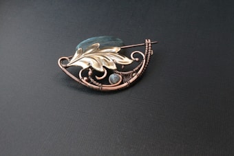 A decorative brooch with intricate wirework, featuring a golden leaf design and a translucent blue stone. The brooch is placed on a dark textured background, highlighting its elegant craftsmanship.