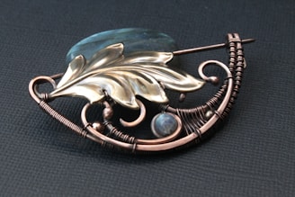 An intricately designed brooch featuring metallic wirework shaped in a curved pattern and adorned with a stylized leaf and bead elements. The wire is crafted in detailed coils and loops, accompanied by a translucent stone, creating an artistic and elegant appearance.