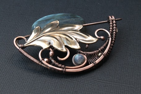 An intricately designed brooch featuring metallic wirework shaped in a curved pattern and adorned with a stylized leaf and bead elements. The wire is crafted in detailed coils and loops, accompanied by a translucent stone, creating an artistic and elegant appearance.