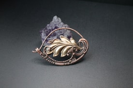 A handcrafted wire-wrapped pendant featuring a gold-toned leaf design intertwined with copper wire. The pendant is placed against a dark textured background, accompanied by a cluster of purple crystals.