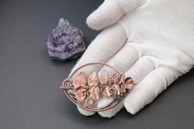 A hand wearing a white glove holds a copper-colored metal piece adorned with intricately designed leaves. Next to the hand, there is a cluster of purple amethyst crystal placed on a dark textured surface.