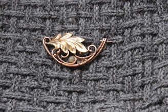 A decorative piece of jewelry featuring a metallic leaf design rests on a textured knit fabric. The brooch is intricately crafted with a combination of copper wires and a small stone accent, creating an elegant and artisanal appearance.