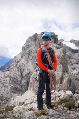 A climber stands confidently on a rocky mountain ledge, wearing a bright orange long-sleeve shirt, blue helmet, and sunglasses. Various climbing gears, including ropes and carabiners, adorn their harness. The rugged, gray cliff face of a mountain looms in the background, with patches of snow visible under a partly cloudy sky.