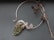 An intricate wire-wrapped piece of jewelry featuring a large green stone is displayed alongside a bare twig with dried seed pods on a dark gray background. The wirework is ornate and elegant, holding the stone in an artistic design.