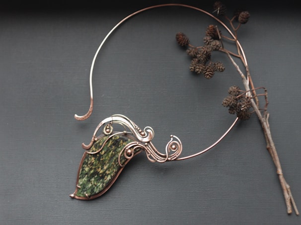An intricate wire-wrapped piece of jewelry featuring a large green stone is displayed alongside a bare twig with dried seed pods on a dark gray background. The wirework is ornate and elegant, holding the stone in an artistic design.