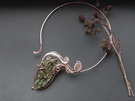 An intricate wire-wrapped piece of jewelry featuring a large green stone is displayed alongside a bare twig with dried seed pods on a dark gray background. The wirework is ornate and elegant, holding the stone in an artistic design.