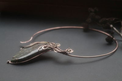An elegant necklace with a unique design featuring a large, polished stone pendant. The stone is encased in intricate wirework that forms swirls and loops, giving it a sophisticated and artistic appearance. The necklace is displayed on a dark surface, and there are blurred floral elements in the background.