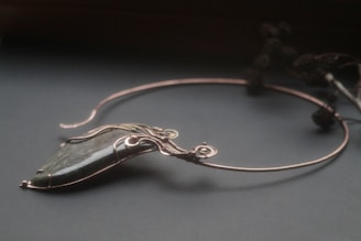 An elegant necklace with a unique design featuring a large, polished stone pendant. The stone is encased in intricate wirework that forms swirls and loops, giving it a sophisticated and artistic appearance. The necklace is displayed on a dark surface, and there are blurred floral elements in the background.